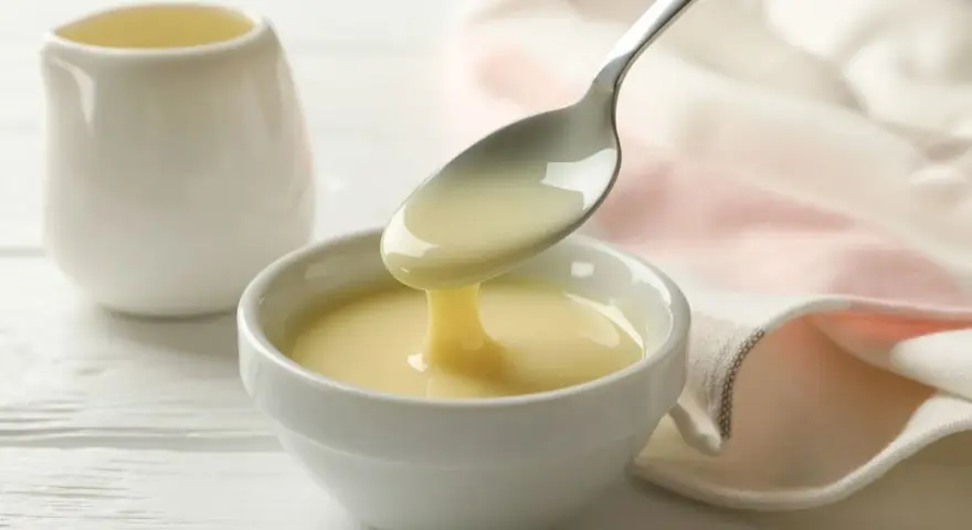 Latte condensato denso e cremoso, perfetto per dolci fatti col Bimby, cola da un cucchiaio in una ciotola bianca.