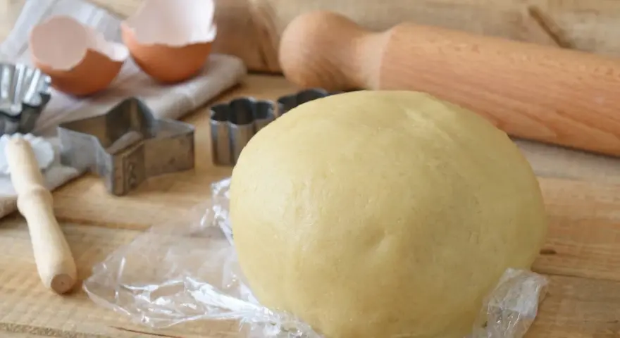 Impasto di pasta frolla con olio, pronto per essere trasformato in biscotti con stampini a forma di stella e fiore. Guscio d'uovo e mattarello completano la scena.
