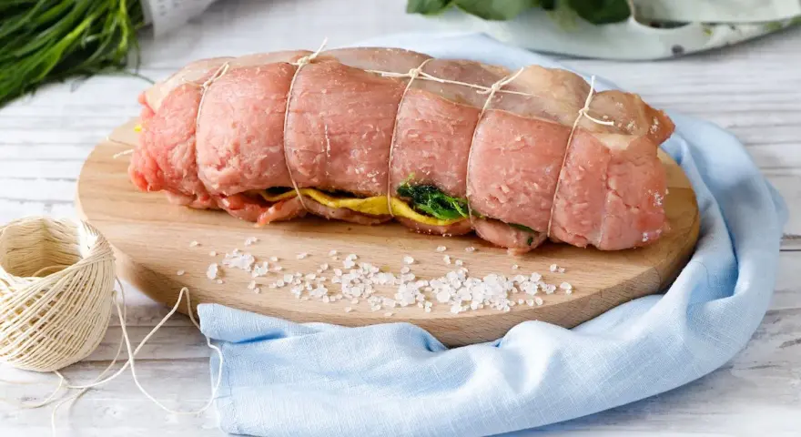 Un arrosto al forno pronto per essere cucinato, legato con spago, su un tagliere con sale grosso e verdure.