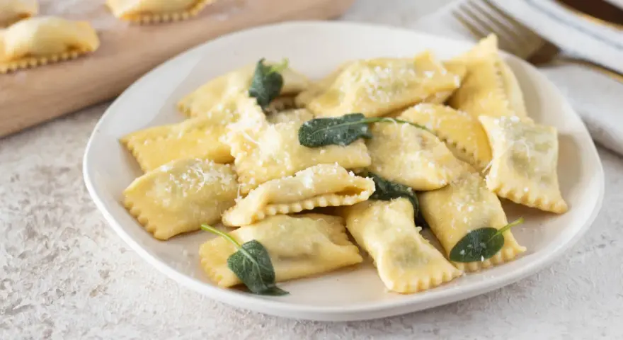 Piatto di ravioli del plin, dorati e spolverati di parmigiano, guarniti con foglie di salvia fresca.