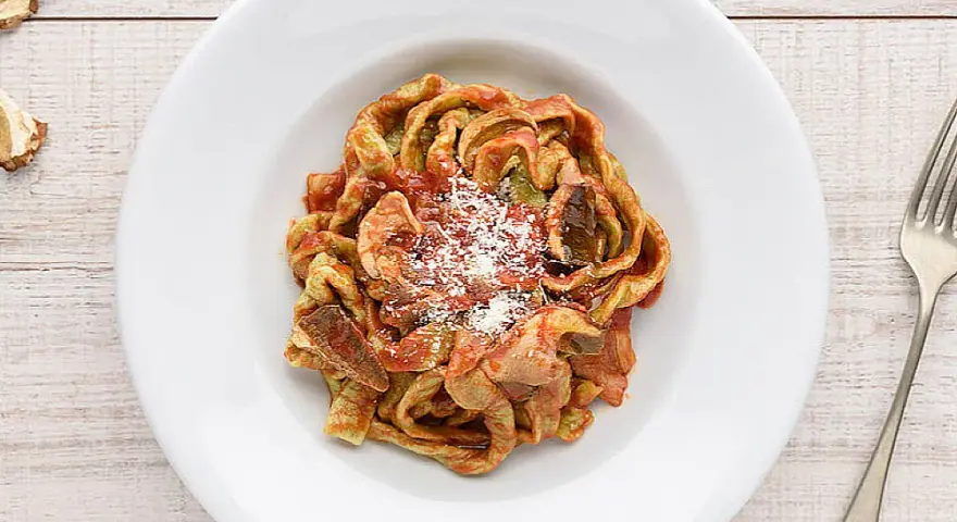 Un piatto di tagliatelle ai funghi, condite con sugo rosso e parmigiano grattugiato, su un tavolo di legno.