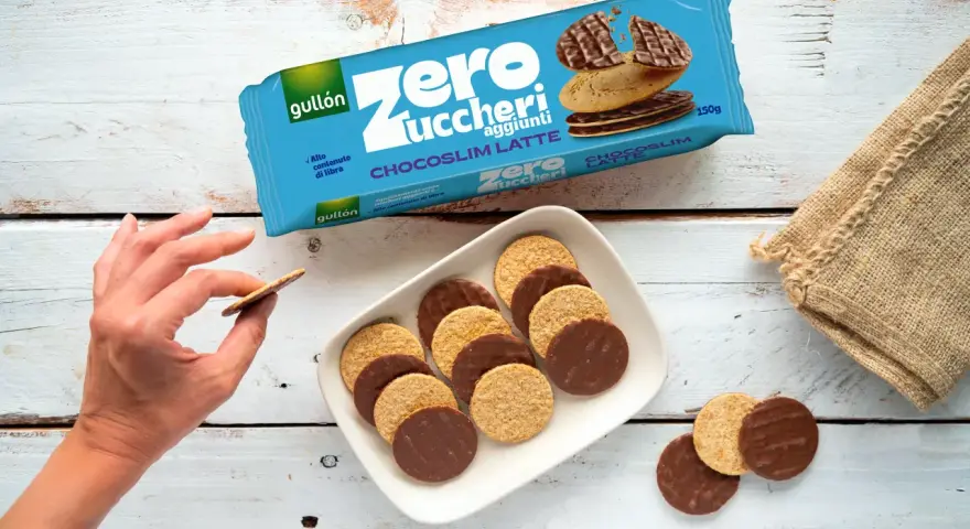 Biscotti senza zucchero Gullón Chocoslim Latte, alcuni ricoperti di cioccolato, su un piatto bianco. Una mano ne prende uno.