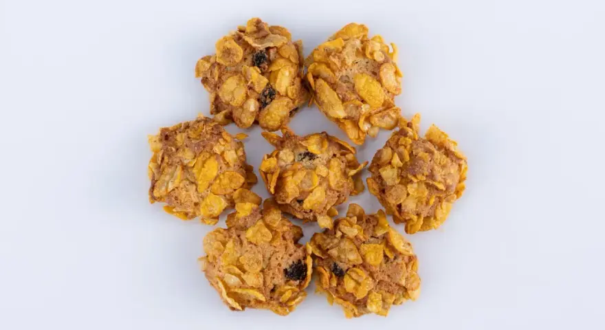 Biscotti croccanti, simili a rose del deserto, con fiocchi di mais e uvetta, disposti a cerchio su sfondo bianco.