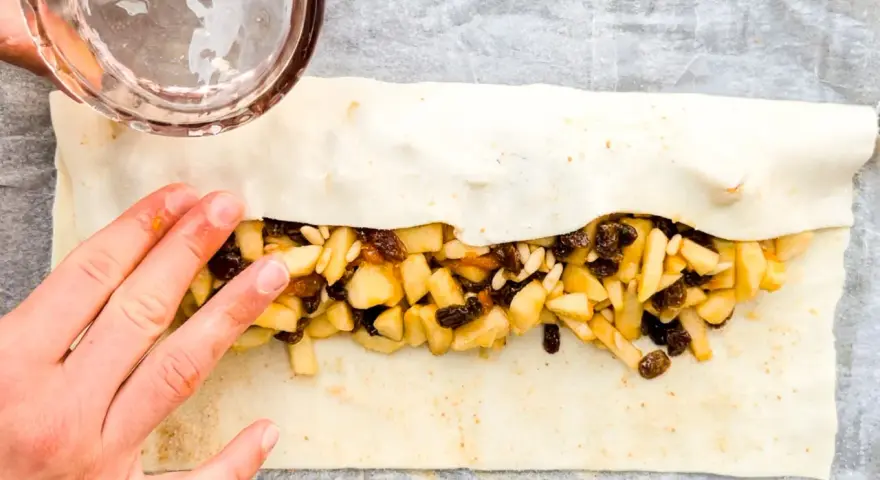 Mani che preparano uno strudel di mele con pasta sfoglia, riempiendolo con mele a cubetti, uvetta e pinoli.
