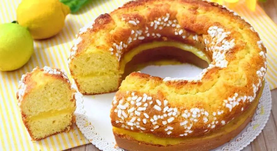 Un soffice ciambellone al limone, decorato con perline di zucchero, con una fetta tagliata che rivela un ripieno cremoso.