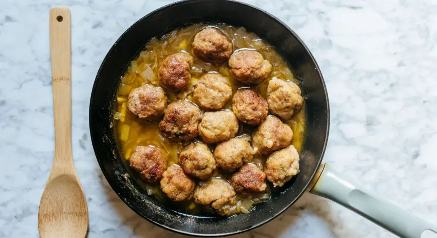 Polpette dorate in padella con cipolle, pronte per essere gustate. Un cucchiaio di legno attende accanto.