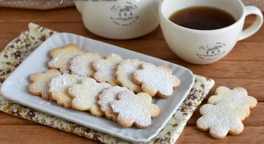 Biscotti con farina di riso a forma di fiore, spolverati di zucchero a velo, accanto a una tazza di tè e una teiera.