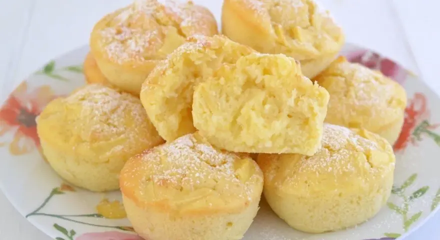 Morbidi muffin alle mele, spolverati di zucchero a velo, su un piatto fiorito. Uno è tagliato a metà, mostrando la soffice mollica.