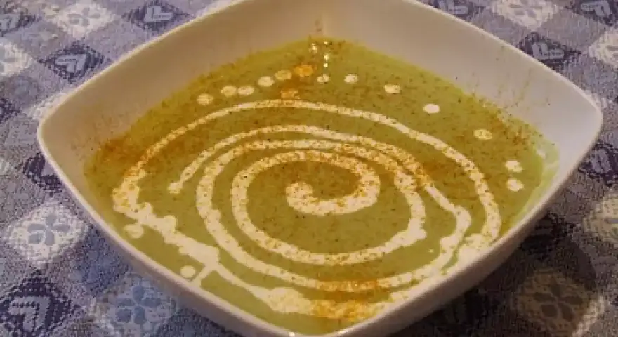 Una ciotola quadrata bianca contiene una vellutata crema di piselli, decorata con un vortice di panna e una spolverata di spezie.