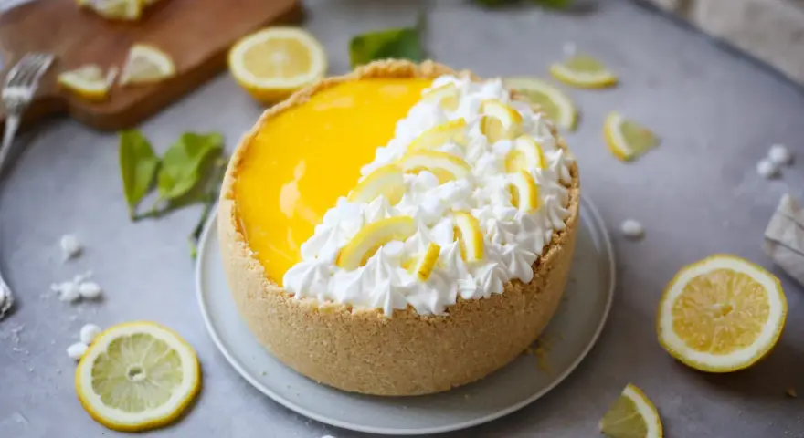 Delizioso cheesecake al limone con panna montata e fettine di limone, circondato da limoni freschi e foglioline verdi.