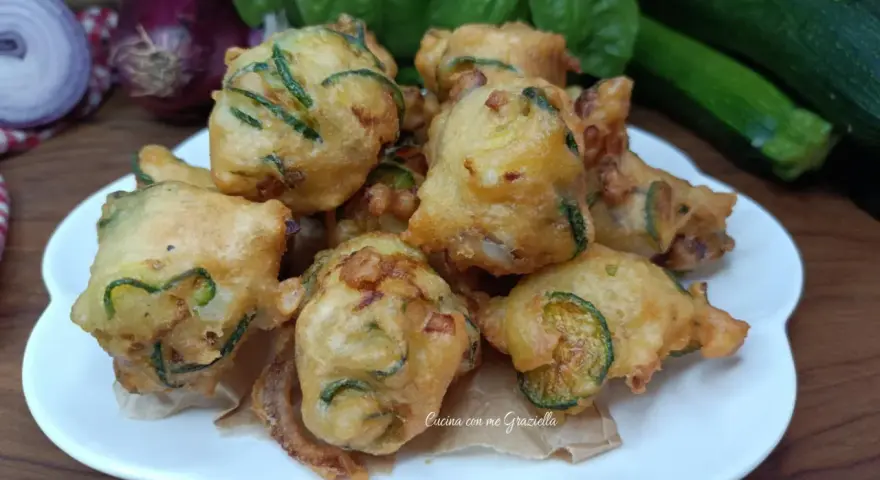 Frittelle di zucchine dorate e croccanti, pronte per essere gustate. Sullo sfondo, cipolle rosse, basilico fresco e zucchine.