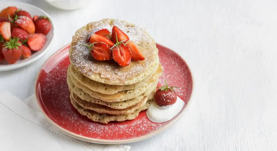 Pila di pancake senza glutine guarniti con fragole fresche e zucchero a velo, serviti su un piatto rosso con un cucchiaio di panna.