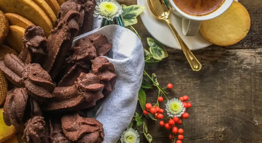 Cesto pieno di biscotti fatti in casa al cioccolato e alla vaniglia, accanto a una tazzina di caffè e bacche rosse.