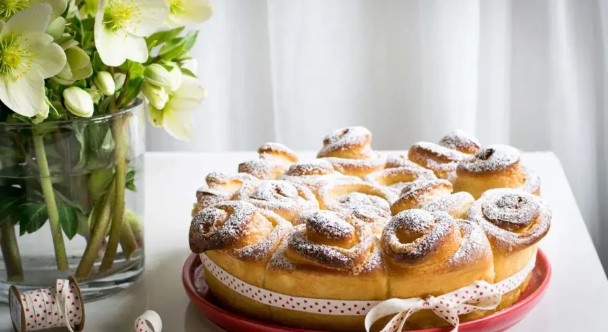 Una deliziosa torta di rose, cosparsa di zucchero a velo, su un piatto rosso, con un nastro a cuoricini. Accanto, un vaso con fiori bianchi.