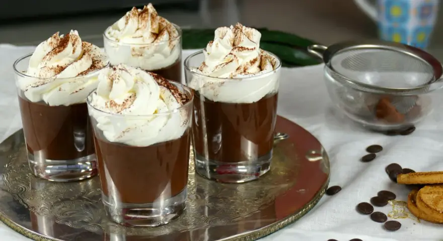 Quattro bicchierini di budino al cioccolato, guarniti con panna montata e cacao in polvere, su un vassoio d'argento.