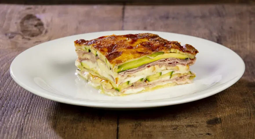 Una fetta di parmigiana di zucchine bianca, stratificata con prosciutto e formaggio filante, su un piatto bianco.