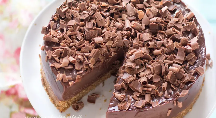 Cheesecake alla Nutella golosa, ricoperta di riccioli di cioccolato fondente. Una fetta è già stata servita, invitando a gustarla.