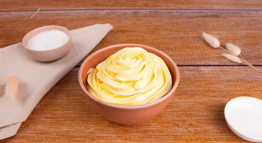 Una ciotola piena di soffice crema al burro, pronta per decorare torte. Accanto, ciotoline con zucchero e decorazioni naturali.