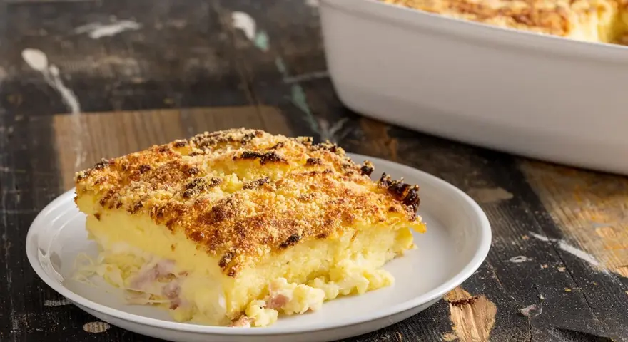Una fetta di sformato di patate dorato e croccante, con prosciutto e formaggio filante, servita su un piatto bianco.