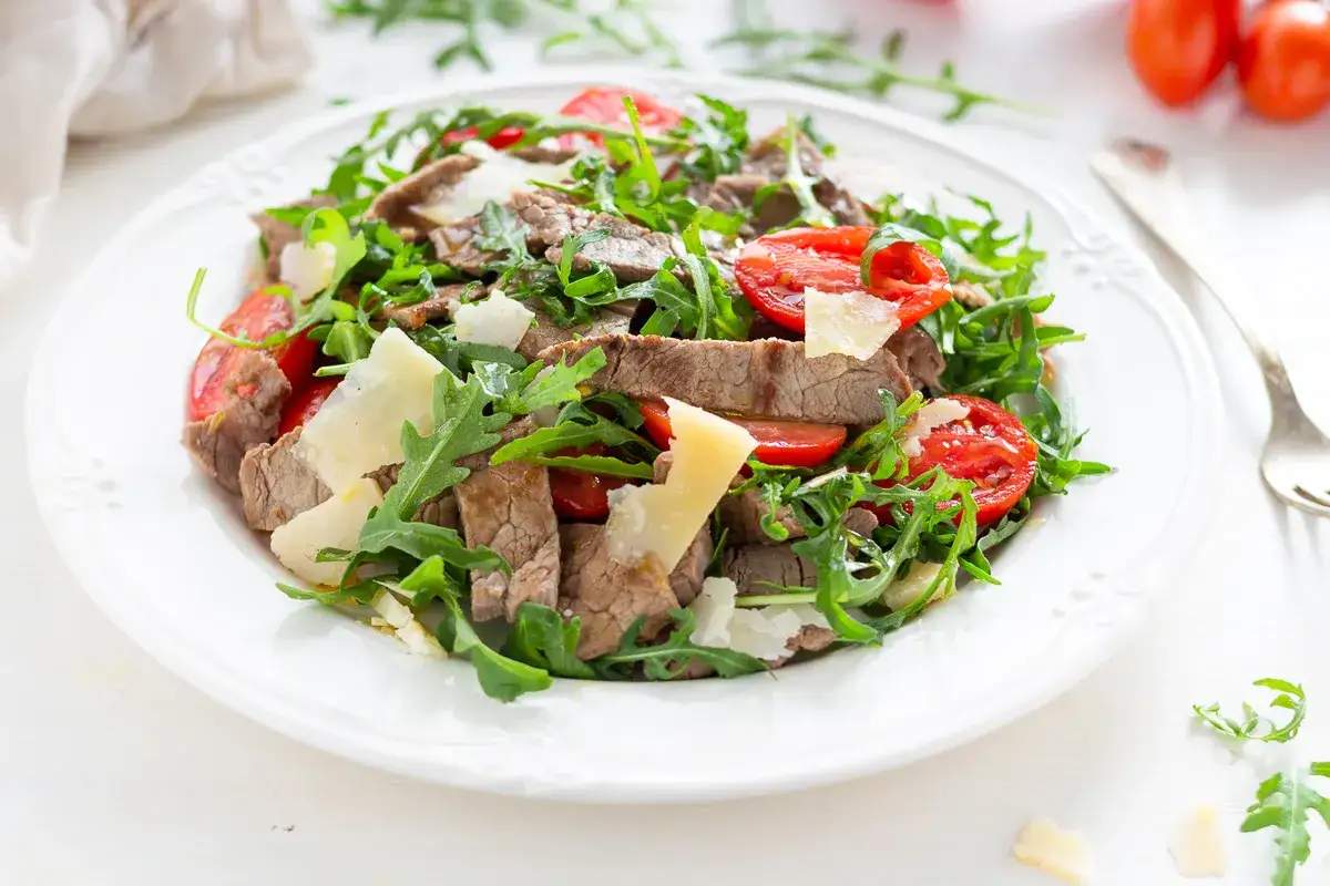 Insalata fresca con straccetti di manzo, pomodorini, rucola e scaglie di parmigiano su un piatto bianco.
