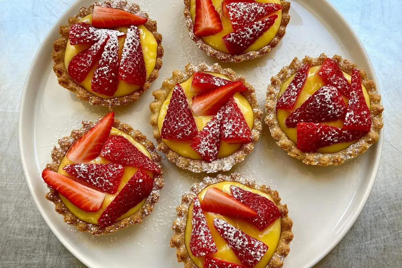 Sei crostatine di fragole con crema gialla, cosparse di zucchero a velo, su un piatto bianco.