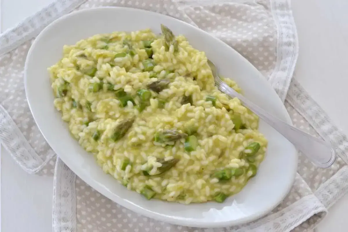 Un cremoso risotto con asparagi, servito su un piatto ovale bianco, con una forchetta d'argento appoggiata sopra.