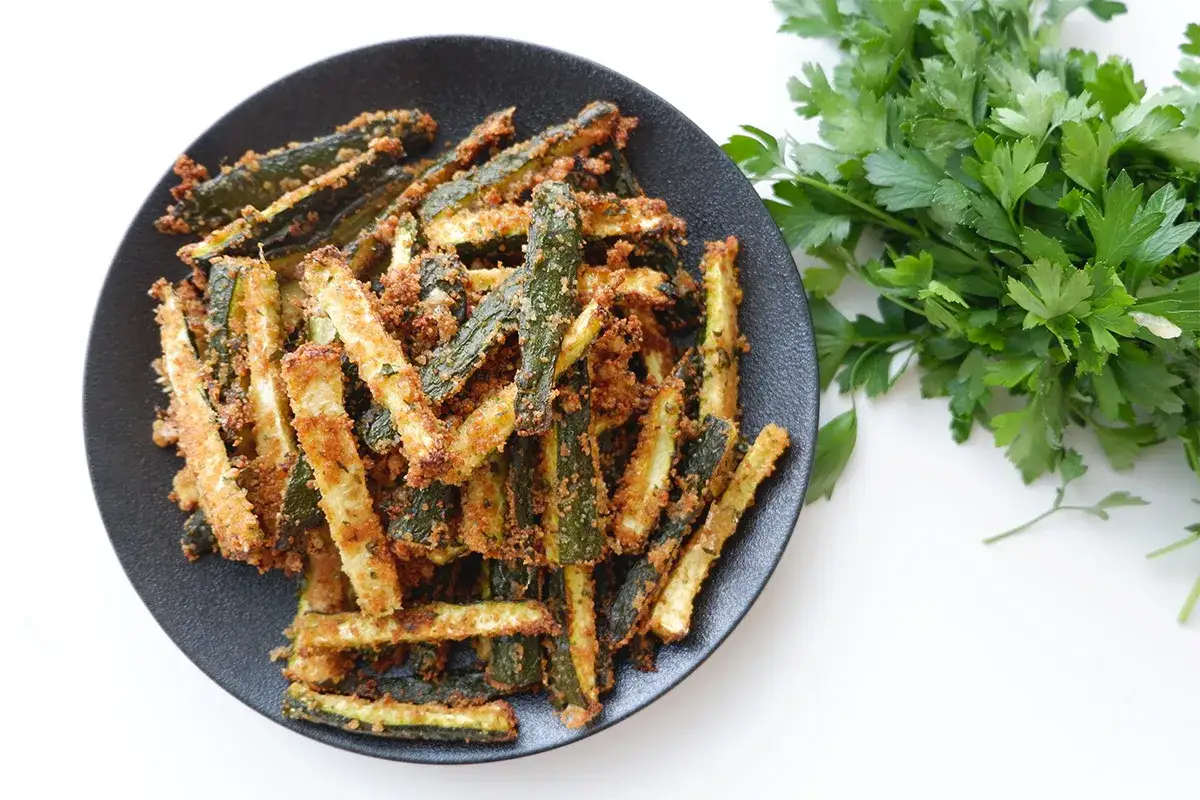 Zucchine gratinate in padella, dorate e croccanti, servite su un piatto nero con un ciuffo di prezzemolo fresco.