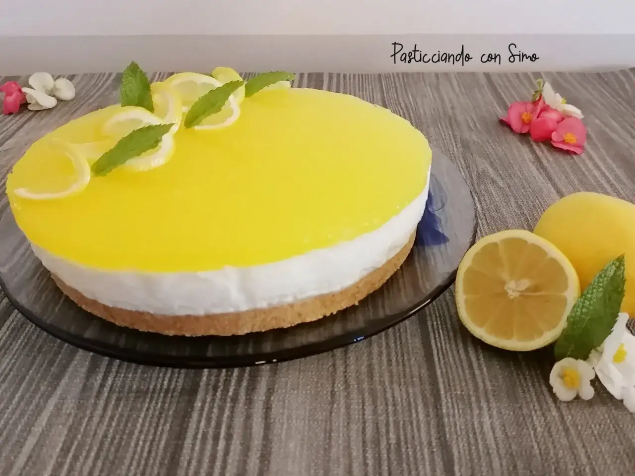 Delizioso cheesecake al limone con decorazioni di fette di limone e menta. Accanto, limoni freschi e fiori rosa e bianchi.
