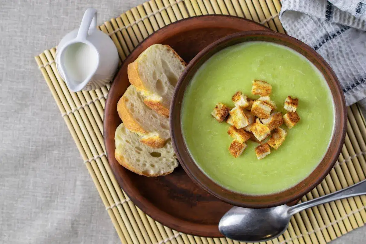 Una ciotola di cremosa crema di zucchine con crostini dorati, accompagnata da fette di pane fresco e una caraffa di latte.