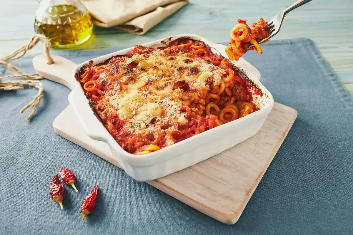 Piatto fumante di pasta alla siciliana, con sugo rosso e formaggio gratinato. Una forchettata mostra la pasta a forma di anello.