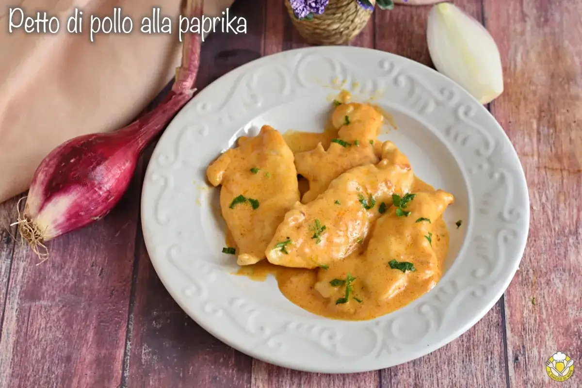Petto di pollo alla paprika, un piatto cremoso e saporito, servito su un piatto bianco con cipolle rosse e bianche sullo sfondo.