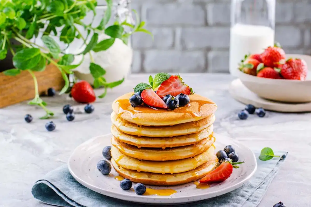 Pancakes senza glutine dorati, impilati e guarniti con fragole, mirtilli e sciroppo. Un piacere per gli occhi e per il palato.