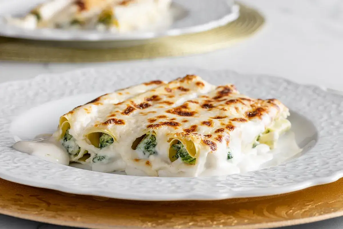 Cannelloni ricotta e spinaci dorati e filanti, serviti su un piatto bianco decorato, con una cremosa besciamella.