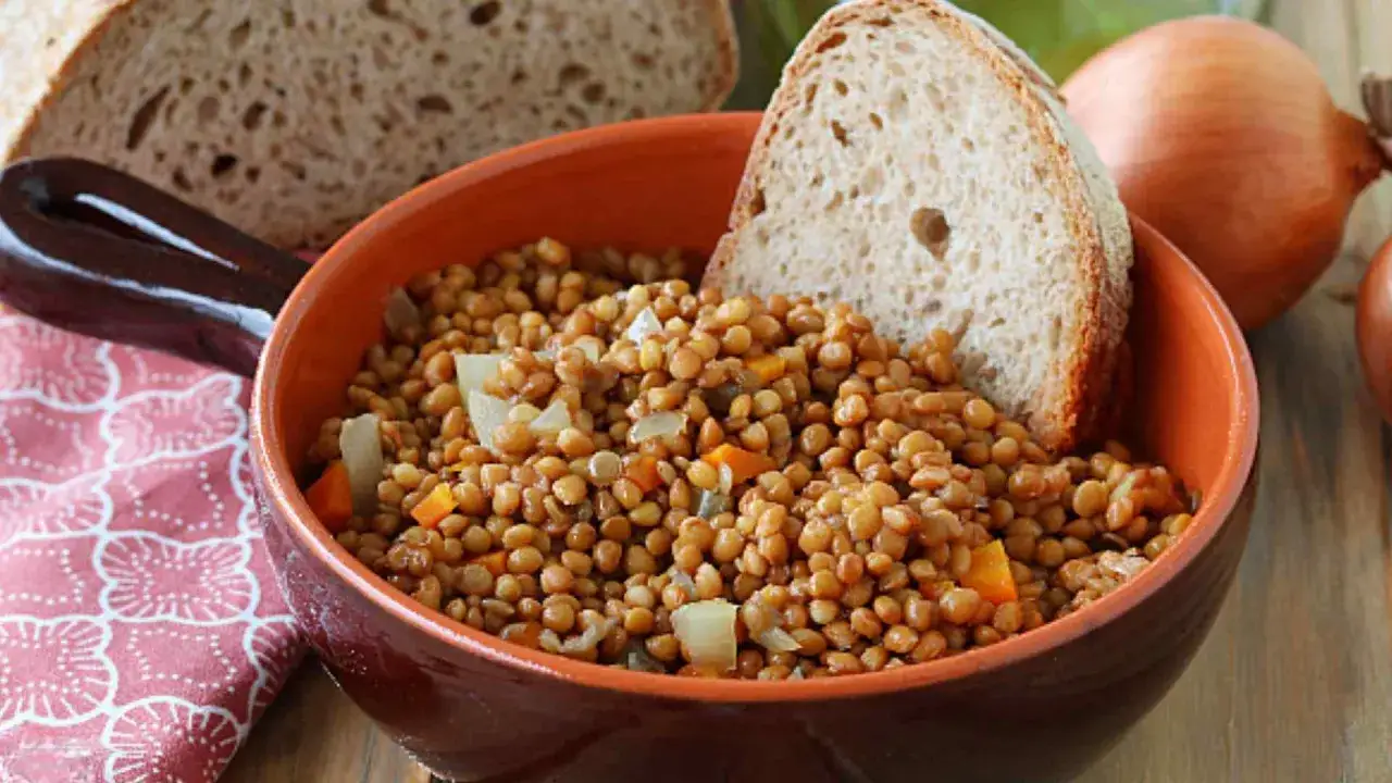 Una ciotola di terracotta piena di lenticchie in umido, con pezzi di carota e cipolla, accompagnata da pane rustico e cipolle intere sullo sfondo.