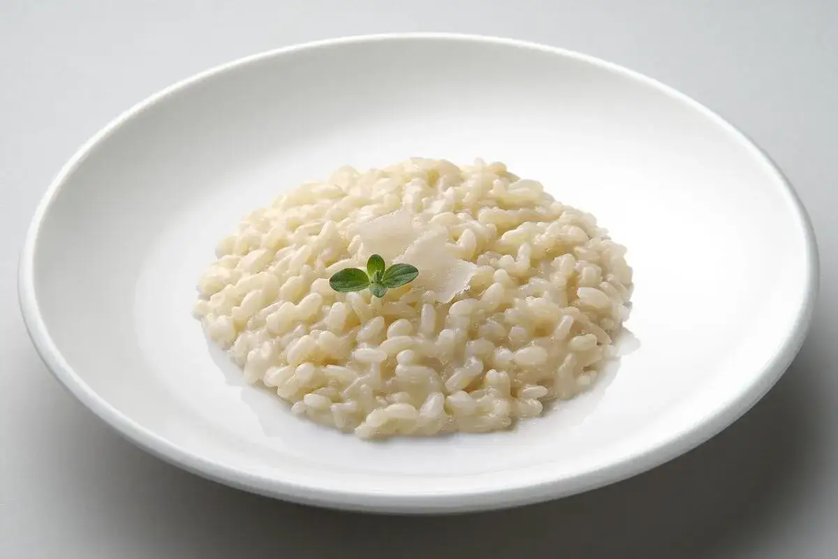 Un cremoso risotto in cagnone, guarnito con scaglie di parmigiano e una fogliolina verde, servito in un piatto bianco.