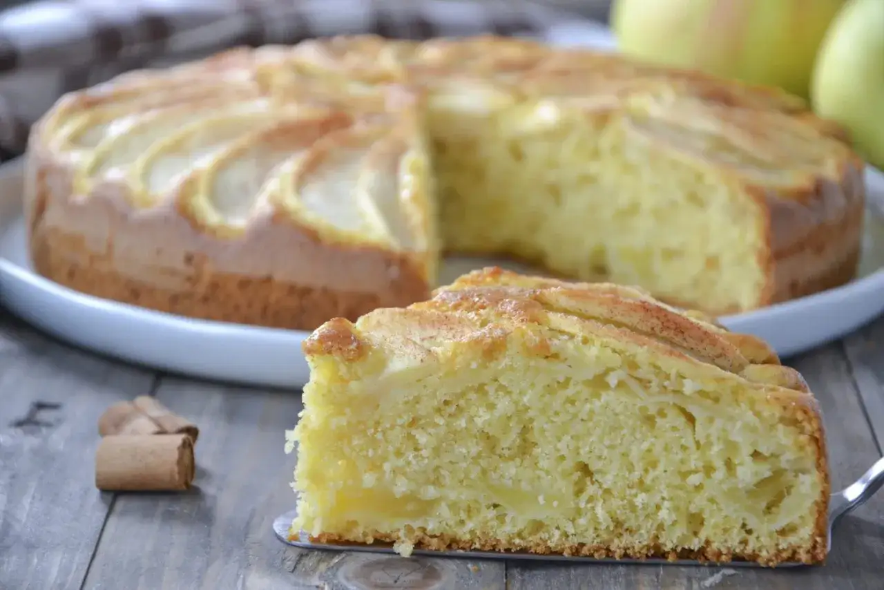 Una fetta di torta di mele senza burro, soffice e profumata, con mele a vista e cannella. La torta intera è sullo sfondo.