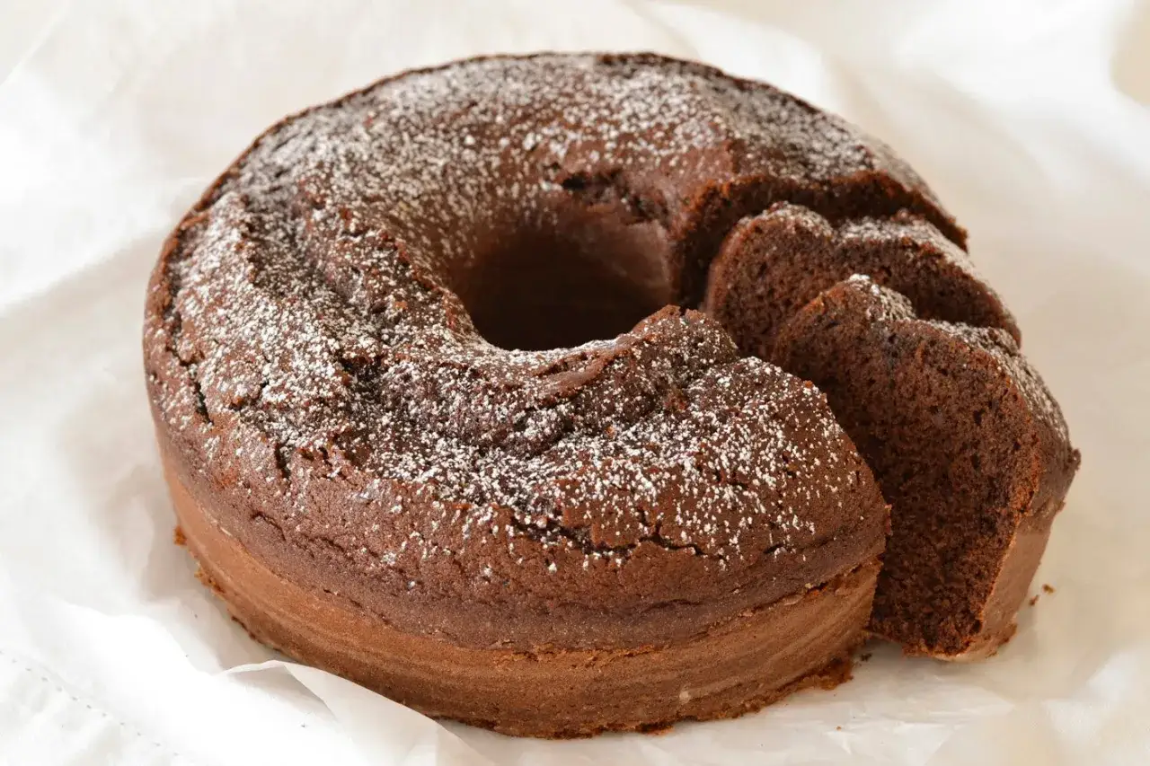 Un soffice ciambellone al cioccolato, cosparso di zucchero a velo, con una fetta tagliata che ne mostra la morbidezza.