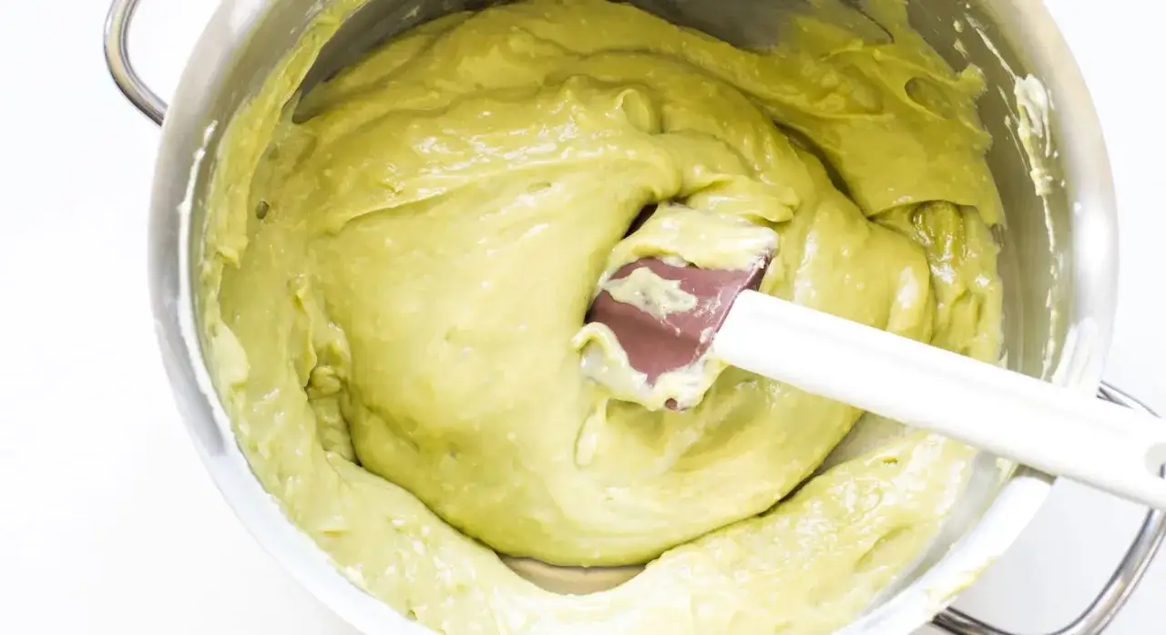 Crema al pistacchio liscia e vellutata in una ciotola d'acciaio, con una spatola che mescola.