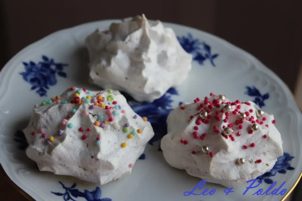 Tre meringhe su un piatto blu e bianco. Due sono decorate con zuccherini colorati e perline argentate. Sono brutti ma buoni, perfetti per una pausa dolce.