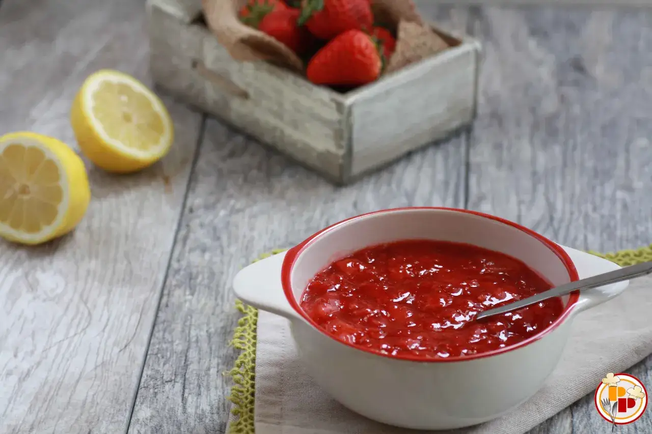 Una ciotola di coulis di fragole, con limoni a fette e fragole fresche in un cesto sullo sfondo.