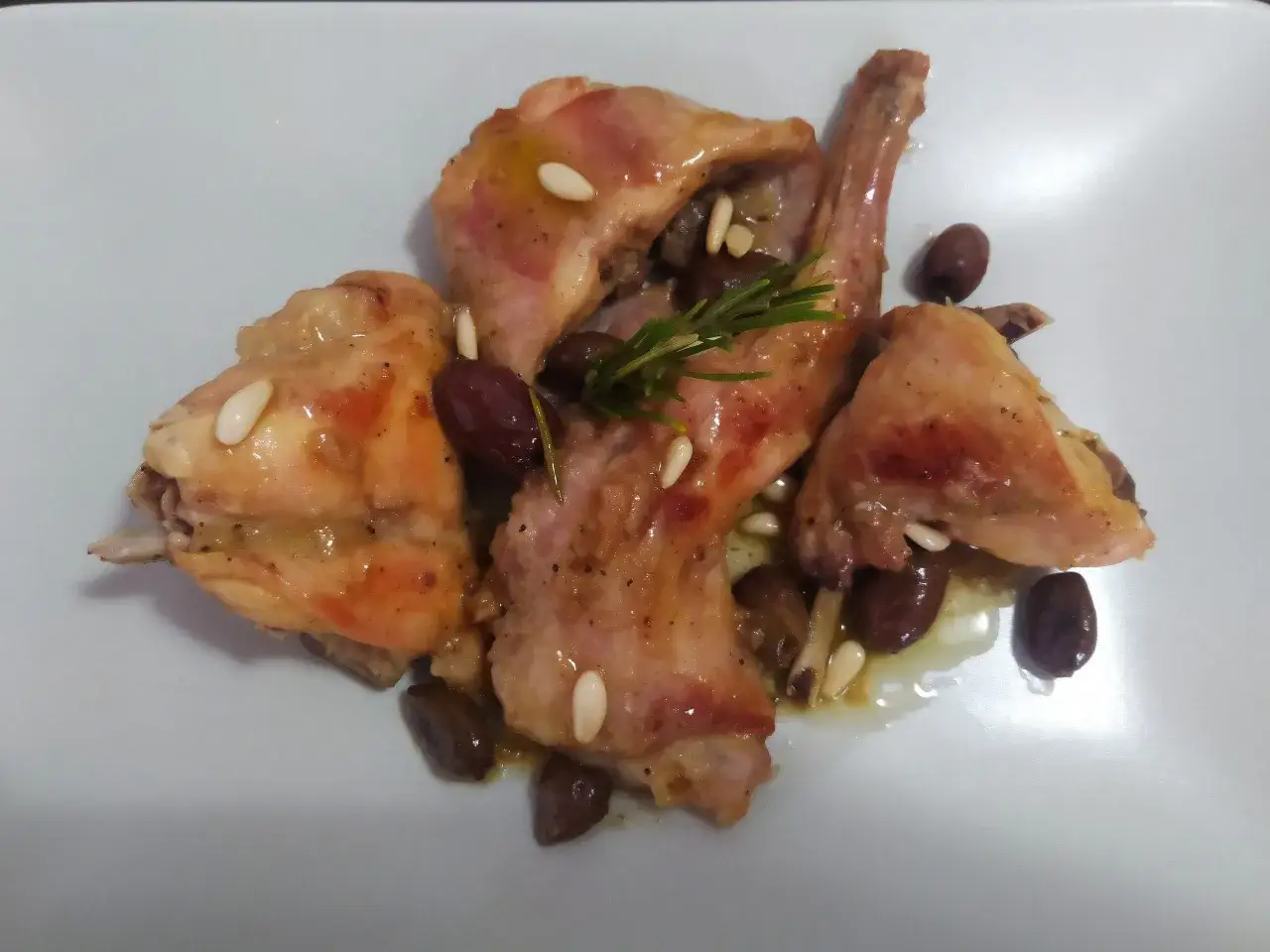 Pezzi di coniglio alla ligure con olive, pinoli e rosmarino su un piatto bianco.