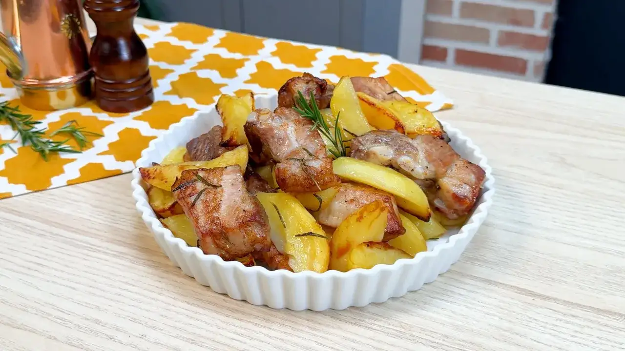 Costine di maiale al forno con patate, profumate al rosmarino, servite in una ciotola bianca.