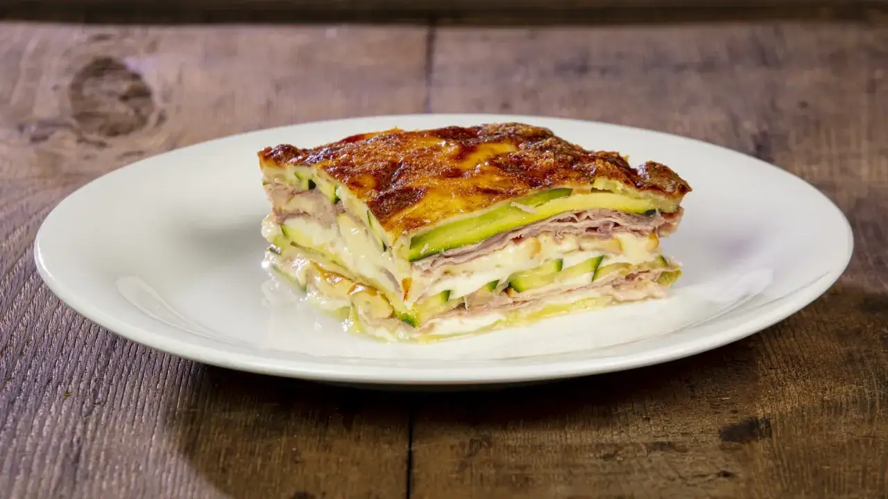 Una fetta di parmigiana di zucchine bianca, stratificata con prosciutto e formaggio fuso, su un piatto bianco.