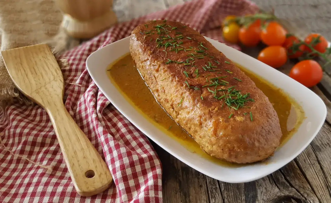 Un succulento polpettone in padella, dorato e profumato, servito su un piatto bianco con erba cipollina. Accanto, una spatola in legno e pomodorini.