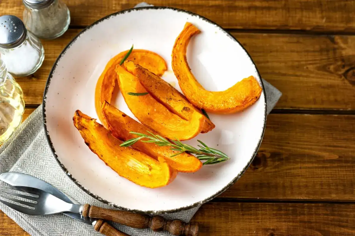 Fette di zucca al forno, dorate e profumate al rosmarino, servite su un piatto bianco con posate in legno.