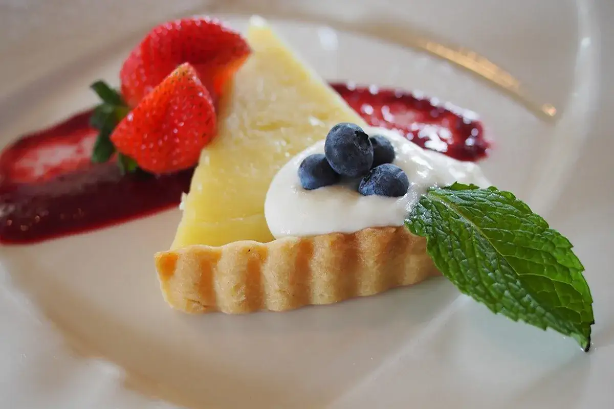 Una fetta di crostata al limone guarnita con panna, mirtilli, fragole e coulis di frutti rossi.
