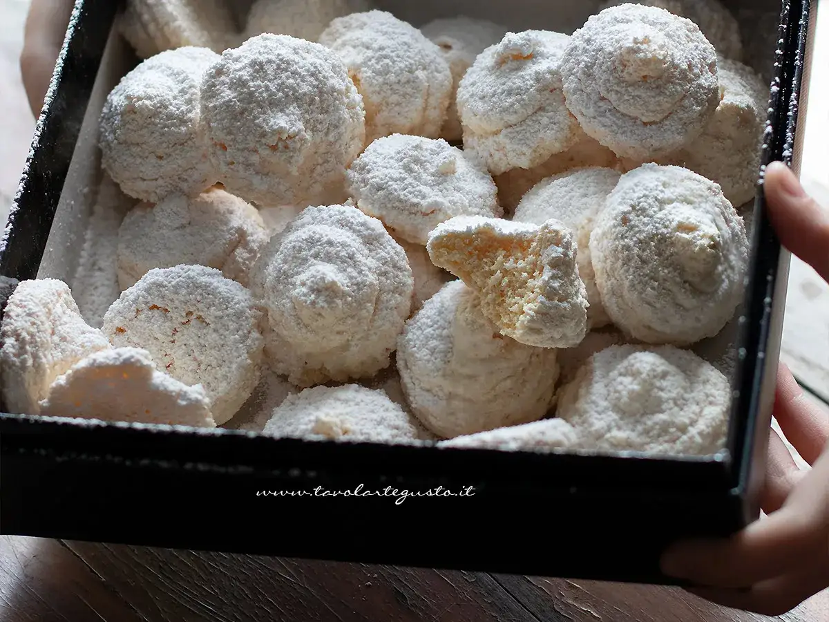 Scatola piena di soffici biscotti al cocco, spolverati di zucchero a velo. Uno &egrave; spezzato, rivelando la sua consistenza friabile.