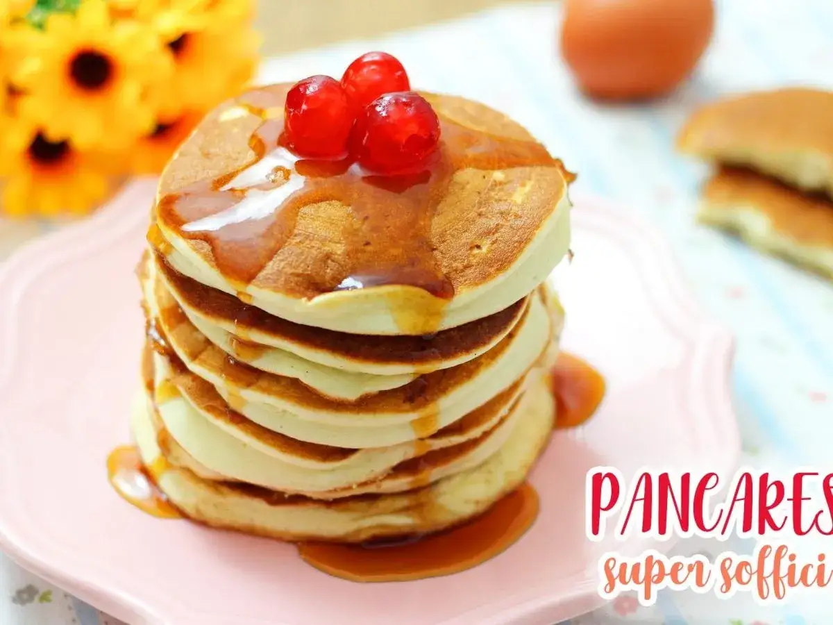 Pancakes senza latte, soffici e dorati, guarniti con sciroppo e ciliegie rosse, su un piatto rosa.