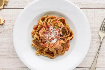 Un piatto di tagliatelle ai funghi, condite con sugo rosso e parmigiano grattugiato, su un tavolo di legno.