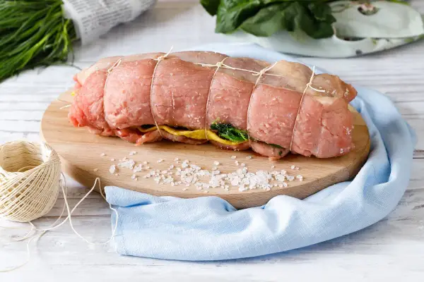 Un arrosto al forno pronto per essere cucinato, legato con spago, su un tagliere con sale grosso e verdure.